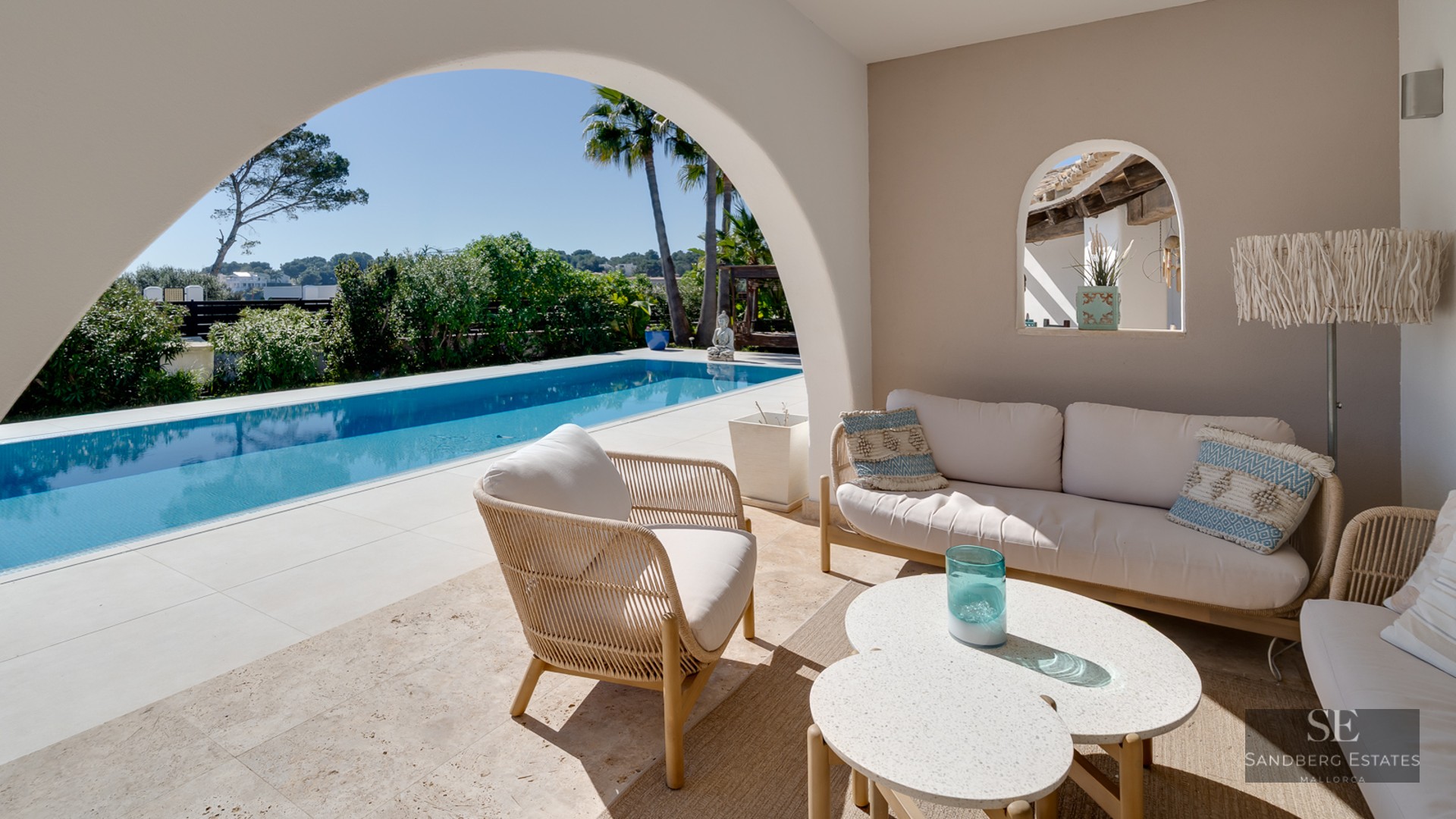 A covered terrace with white lounge furniture looking out through a large archway to a blue swimming pool.