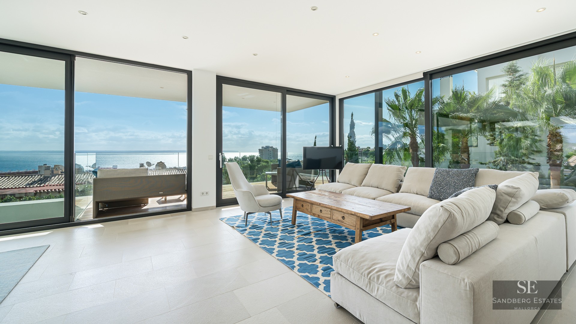Bright living room with beige sofas, a wooden coffee table, and large glass windows overlooking the sea and palm trees.