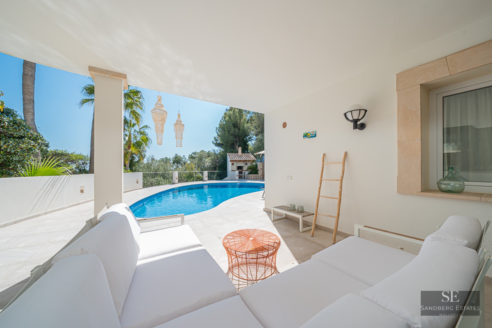 Une élégante terrasse couverte avec des canapés blancs donnant sur une piscine courbe entourée de verdure.