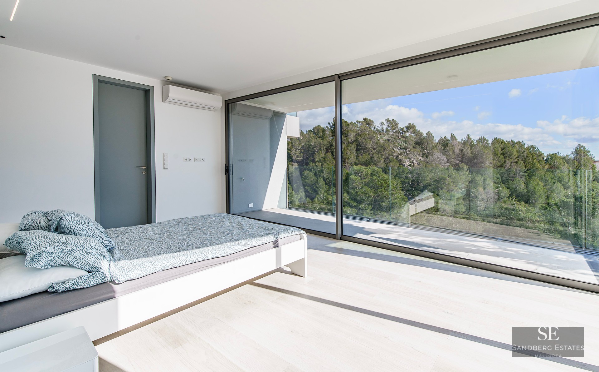 Bright bedroom with floor-to-ceiling glass doors opening to a balcony overlooking a lush green forest.