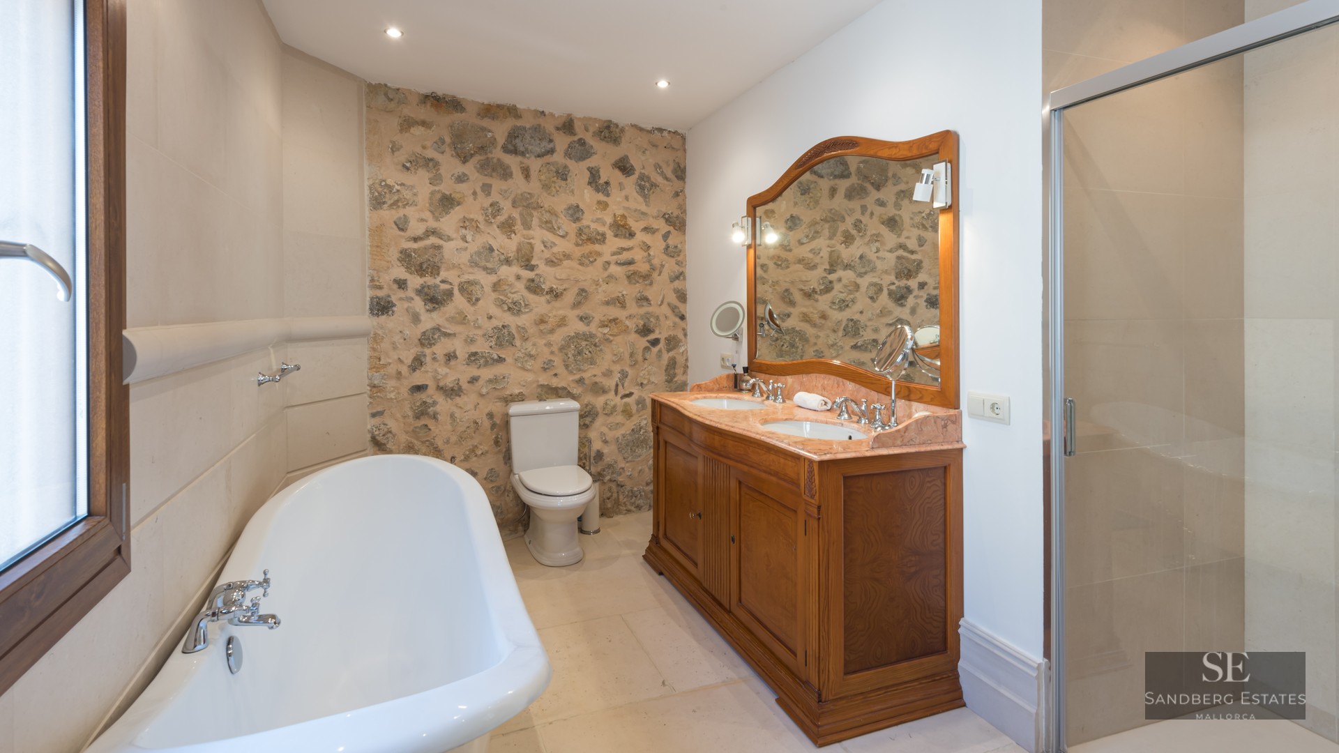 Elegant bathroom with a rustic stone wall, double wooden vanity, freestanding tub, and glass shower enclosure.