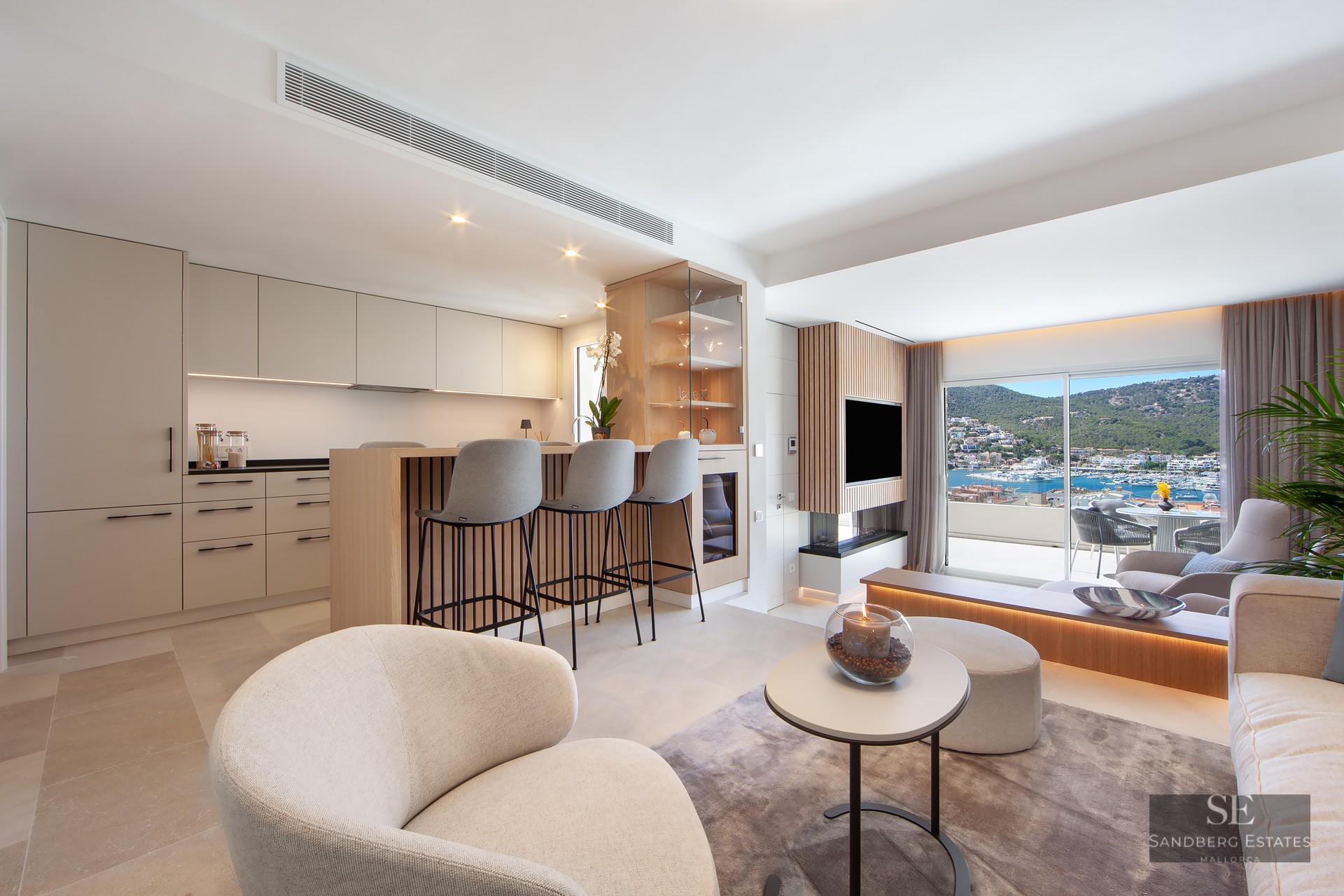 A bright, modern open-plan living room and kitchen featuring beige cabinetry, an island with stools, and a large window with a marina view.