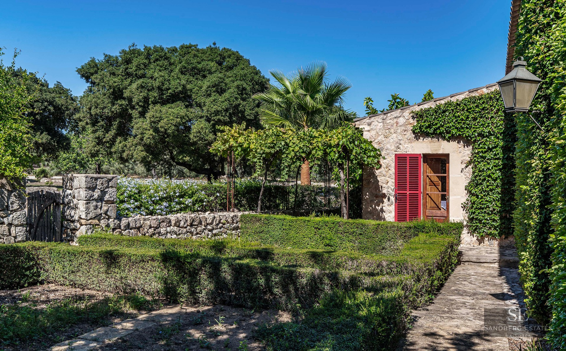 Manicured hedges and stone walls leading to a rustic stone house with red shutters and lush greenery under a blue sky.