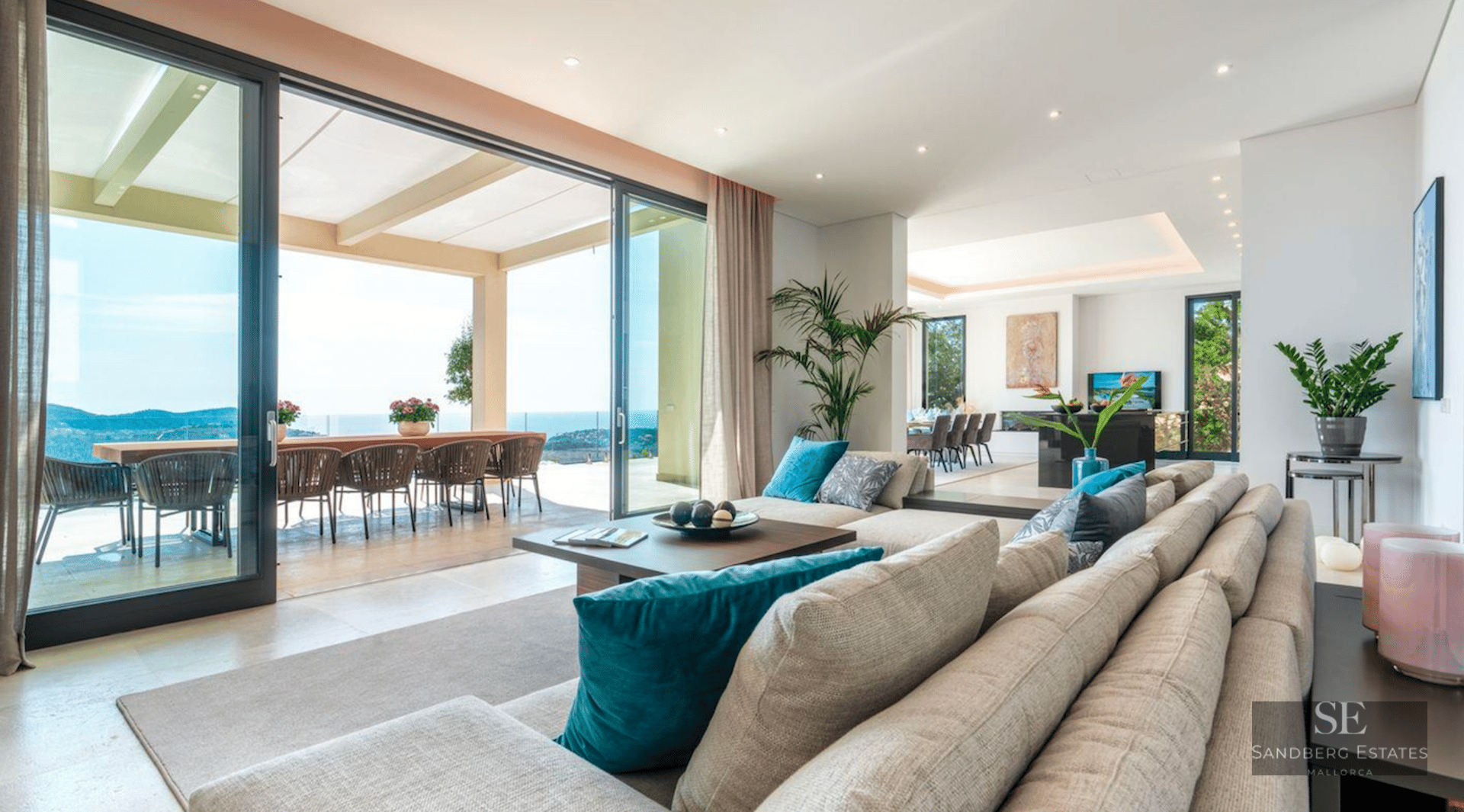 Spacious modern living room with large beige sofa, floor-to-ceiling glass doors opening to a terrace with sea views.