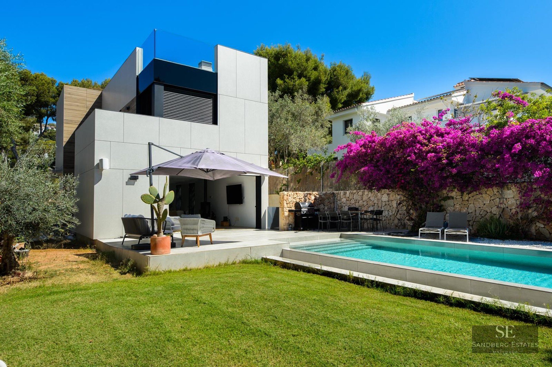Modern villa exterior with a blue rectangular pool, green lawn, and bright purple bougainvillea against a stone wall.