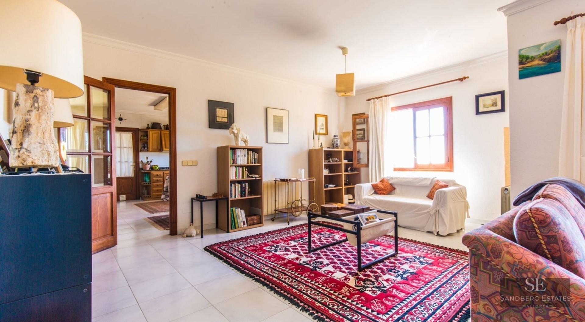 Bright living room with white sofas, red patterned rug, bookshelves, and wood accents leading to a kitchen area.