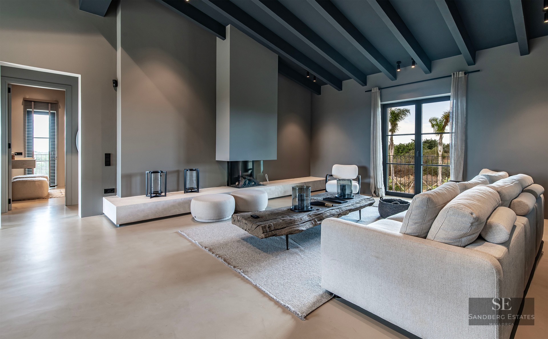 Spacious modern living room with grey ceiling beams, beige sofa, rustic wood table, and built-in minimalist fireplace.