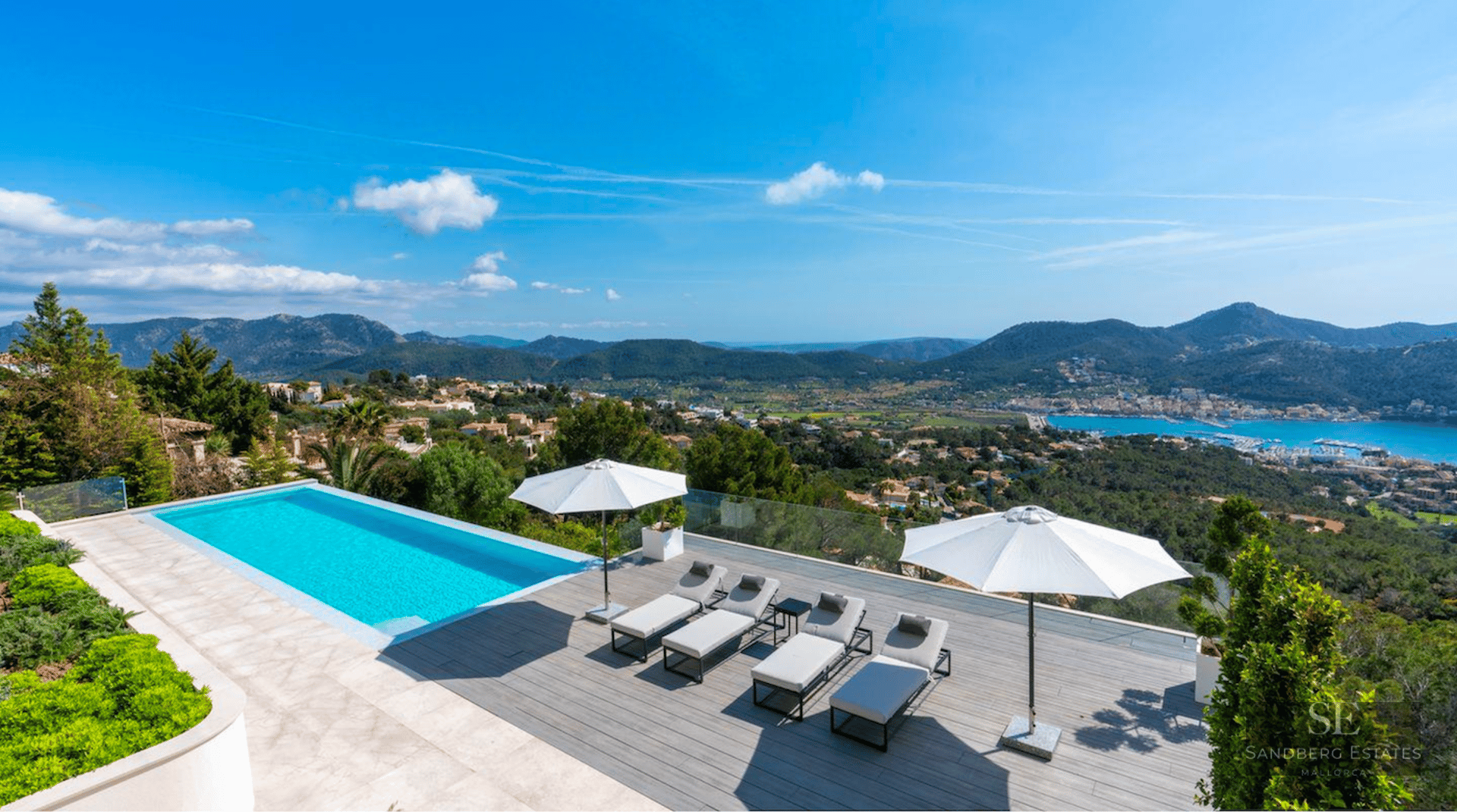 Outdoor terrace featuring a blue swimming pool, modern loungers, white umbrellas, and panoramic mountain and sea views.