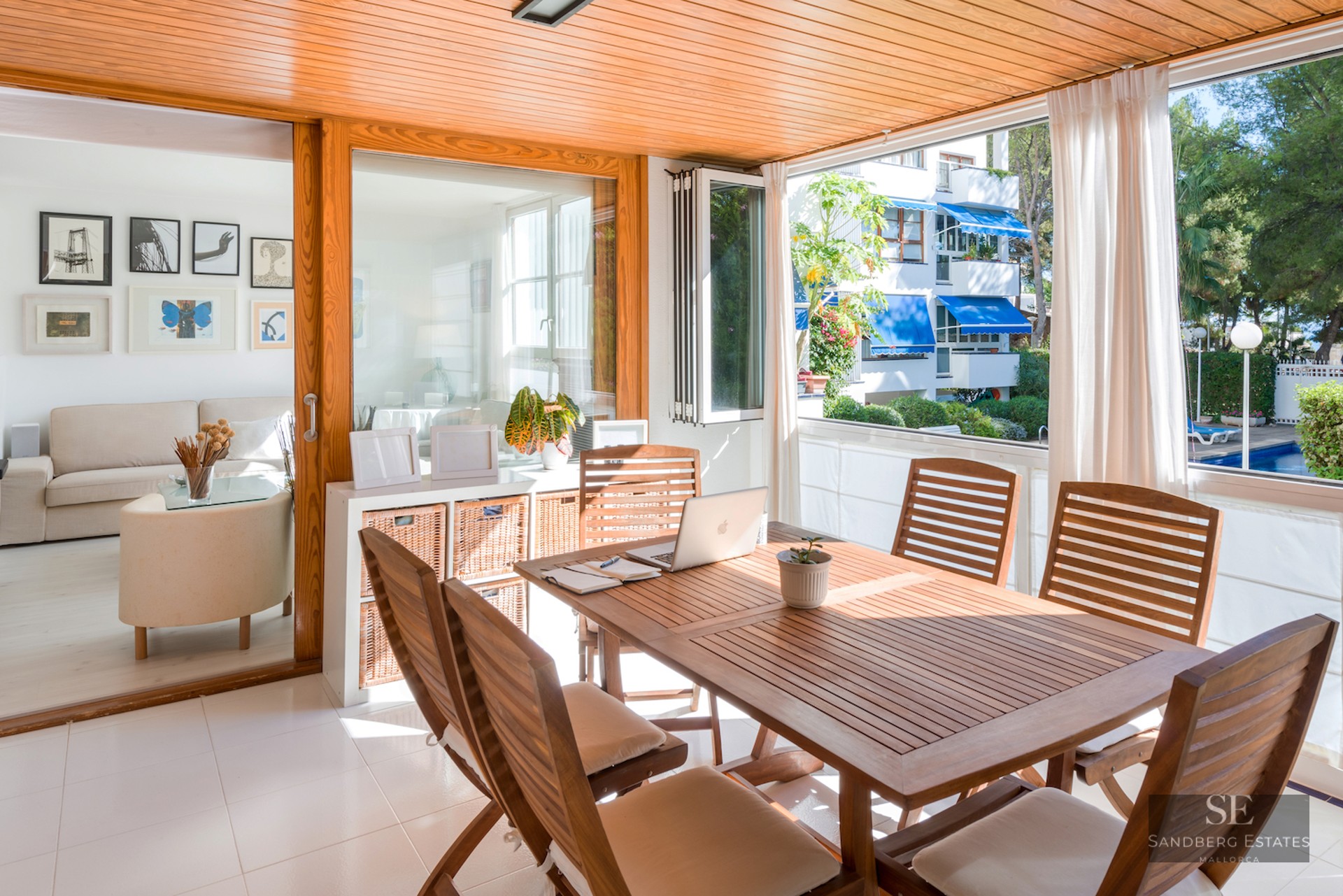 Bright enclosed wooden terrace with dining table and laptop, overlooking a pool and garden through large windows.