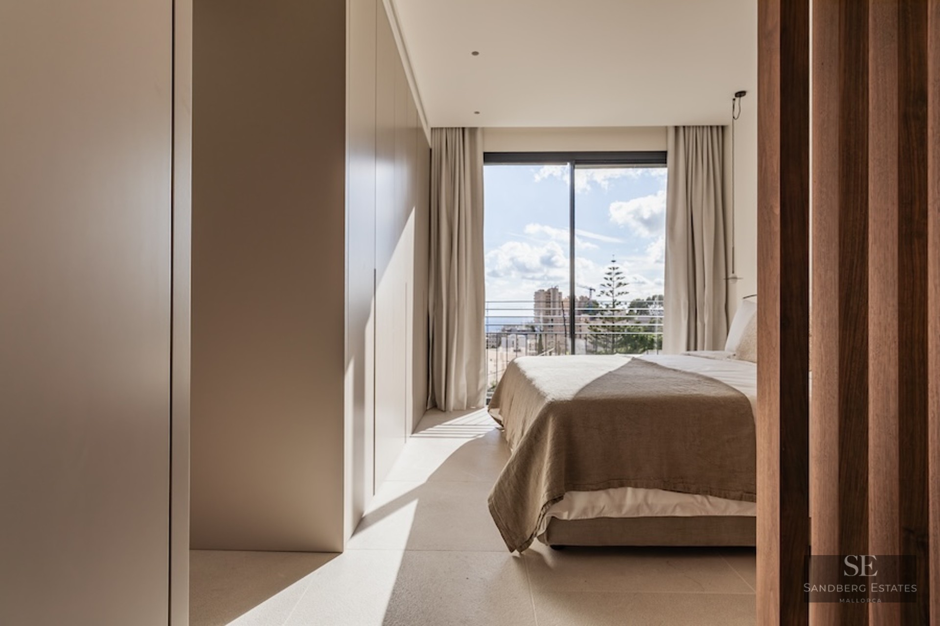 Modern bedroom with beige bedding, built-in wardrobes, and a large sliding glass door opening to a city balcony.