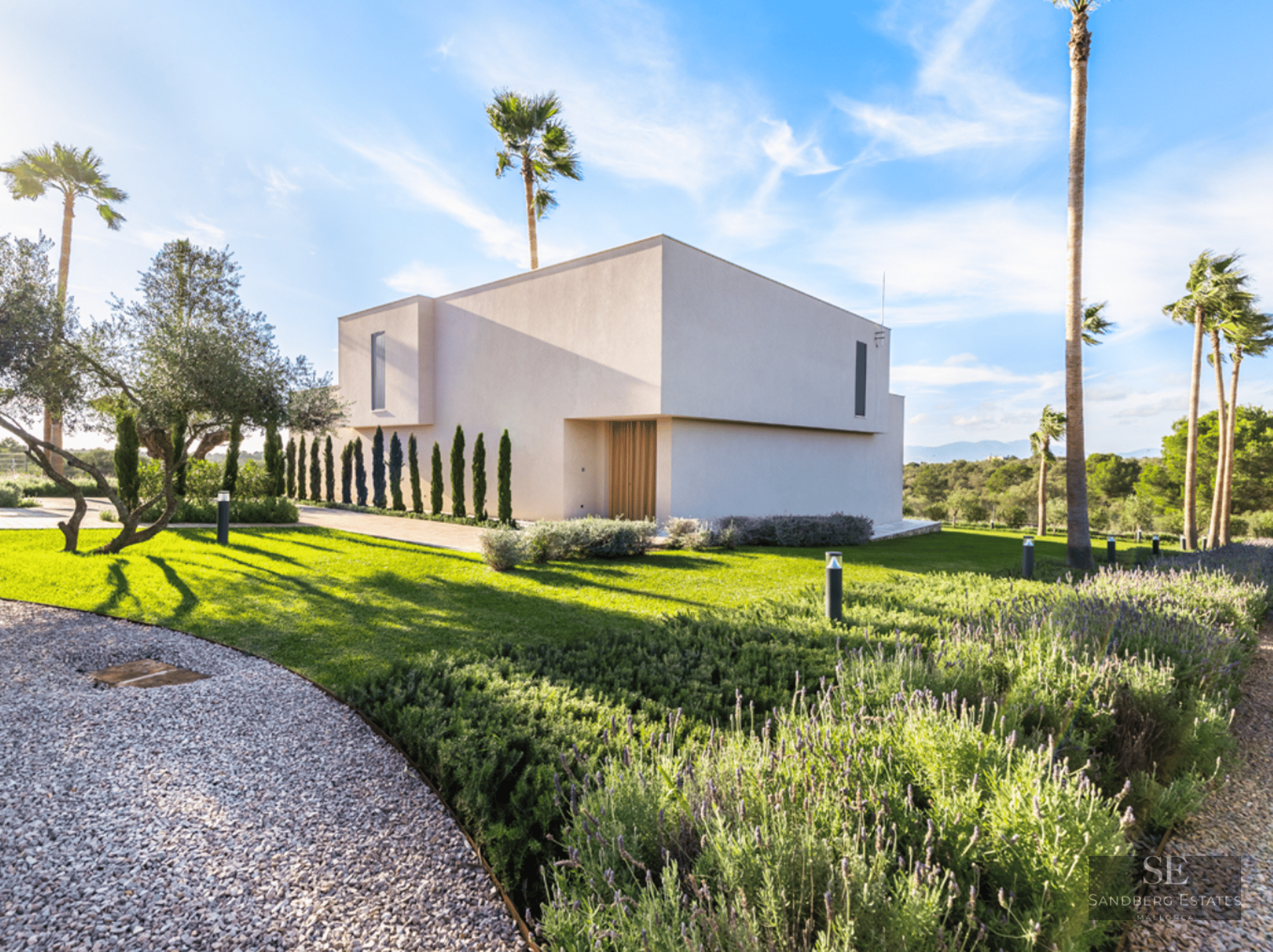 Exterior of a white minimalist villa featuring lavender gardens, green lawn, and palm trees under a bright blue sky.