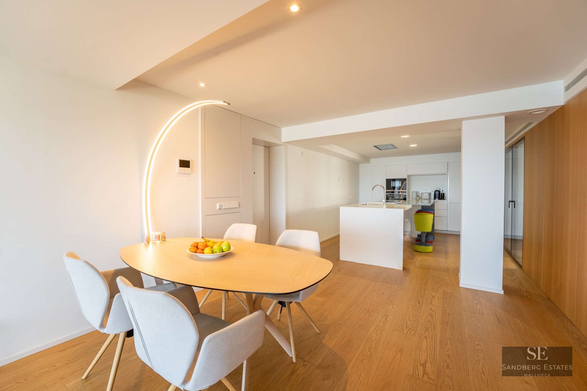 Minimalist dining area with a light wood table, grey chairs, and a striking arched floor lamp, leading to a modern kitchen.