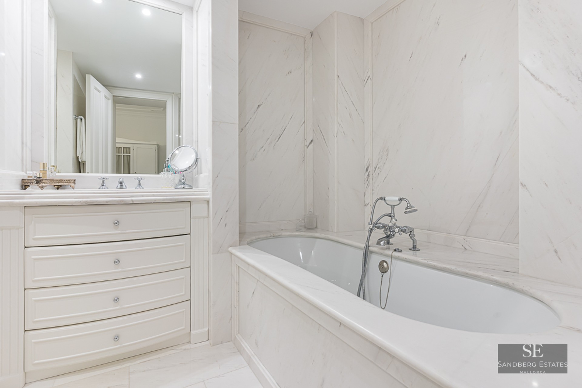 Bright master bathroom featuring white marble walls, a built-in tub with chrome faucets, and a white vanity with drawers.
