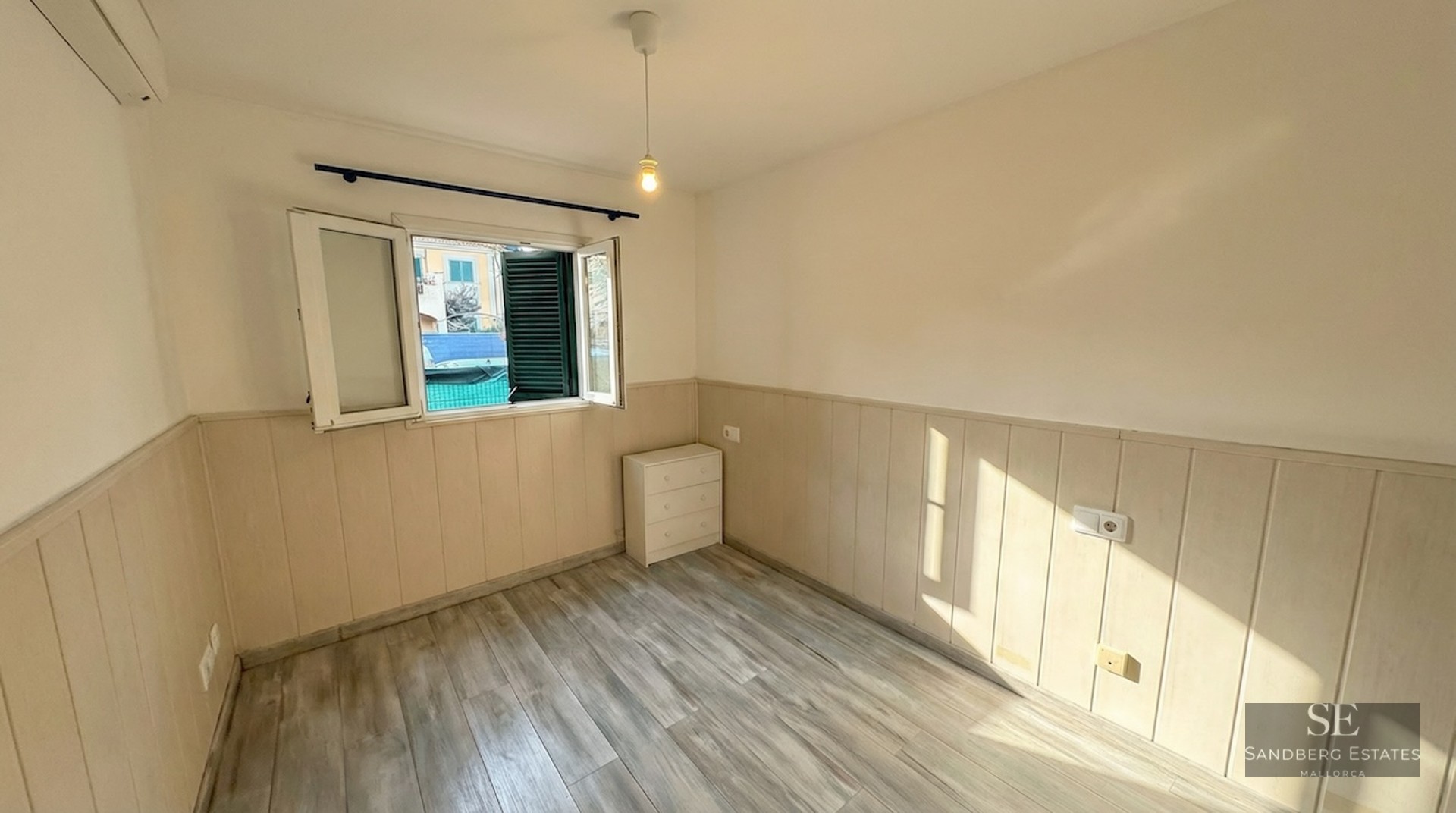 Empty room with light wood floors, partial wall paneling, an open window with green shutters, and a small white dresser.