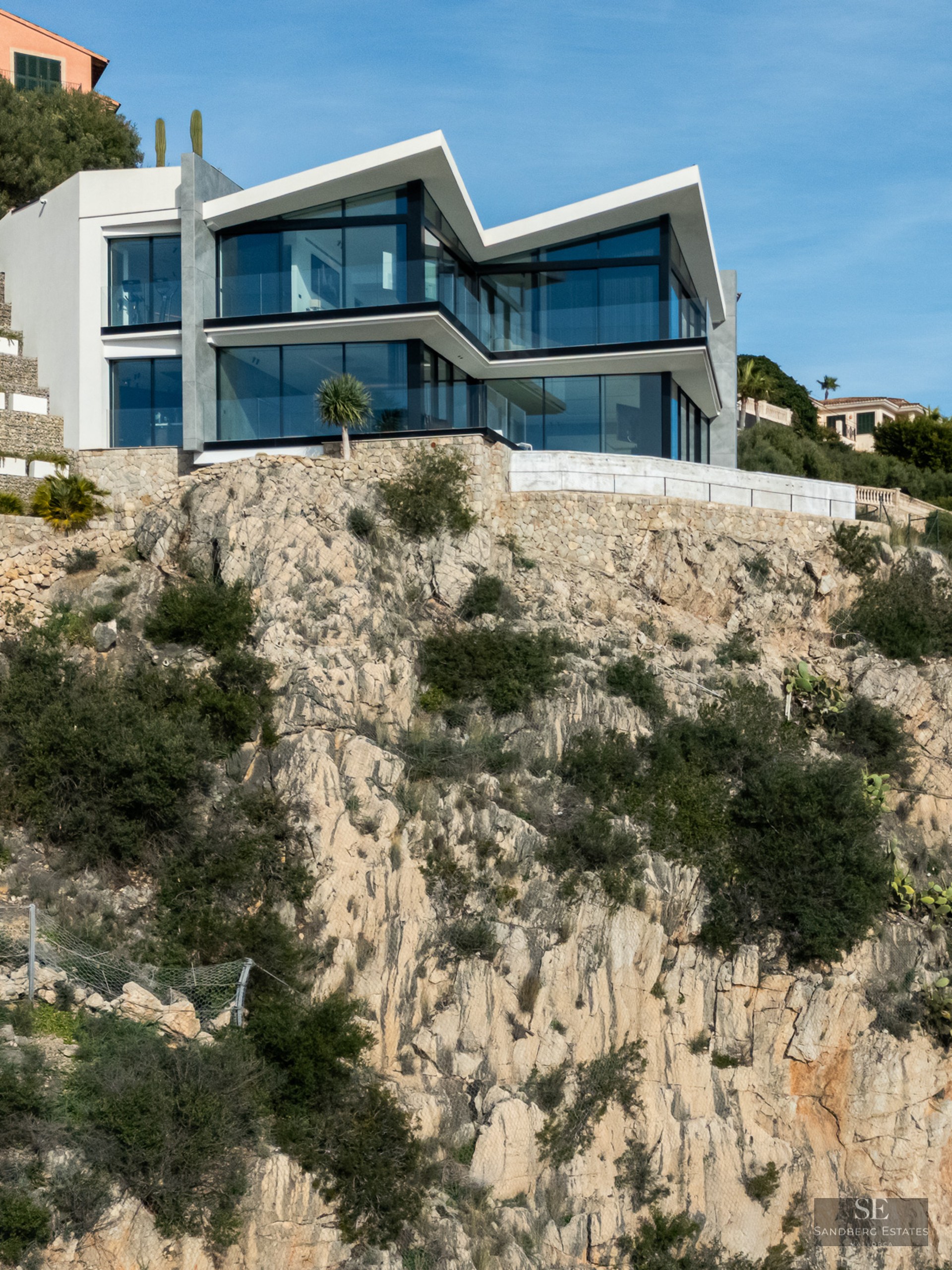 Modern white villa with large glass walls built on a steep rocky cliffside under a clear blue sky.