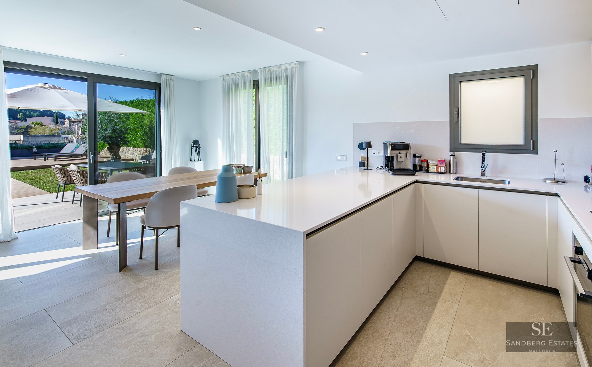 Minimalist white kitchen featuring a large island, wooden dining table, and glass sliding doors leading to a sunny garden.