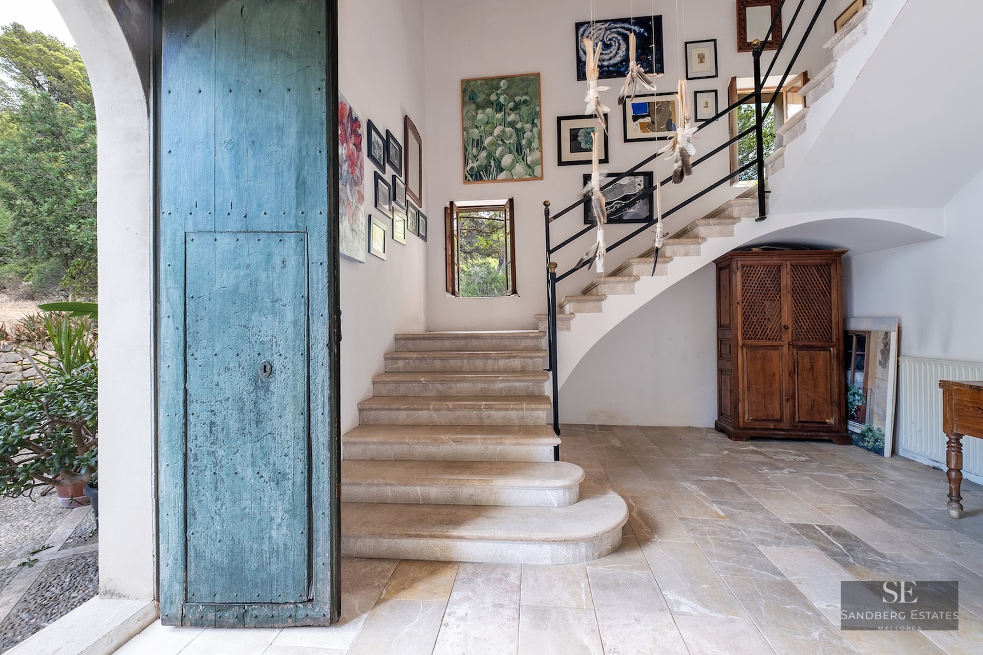 Entrance hall with a large distressed blue door, stone stairs leading up, and a gallery of paintings on white walls.