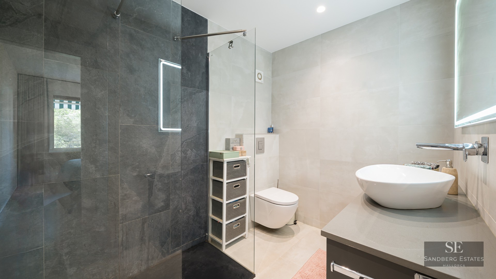 Salle de bain moderne avec douche en verre, carrelage gris foncé, vasque blanche et toilettes suspendues.