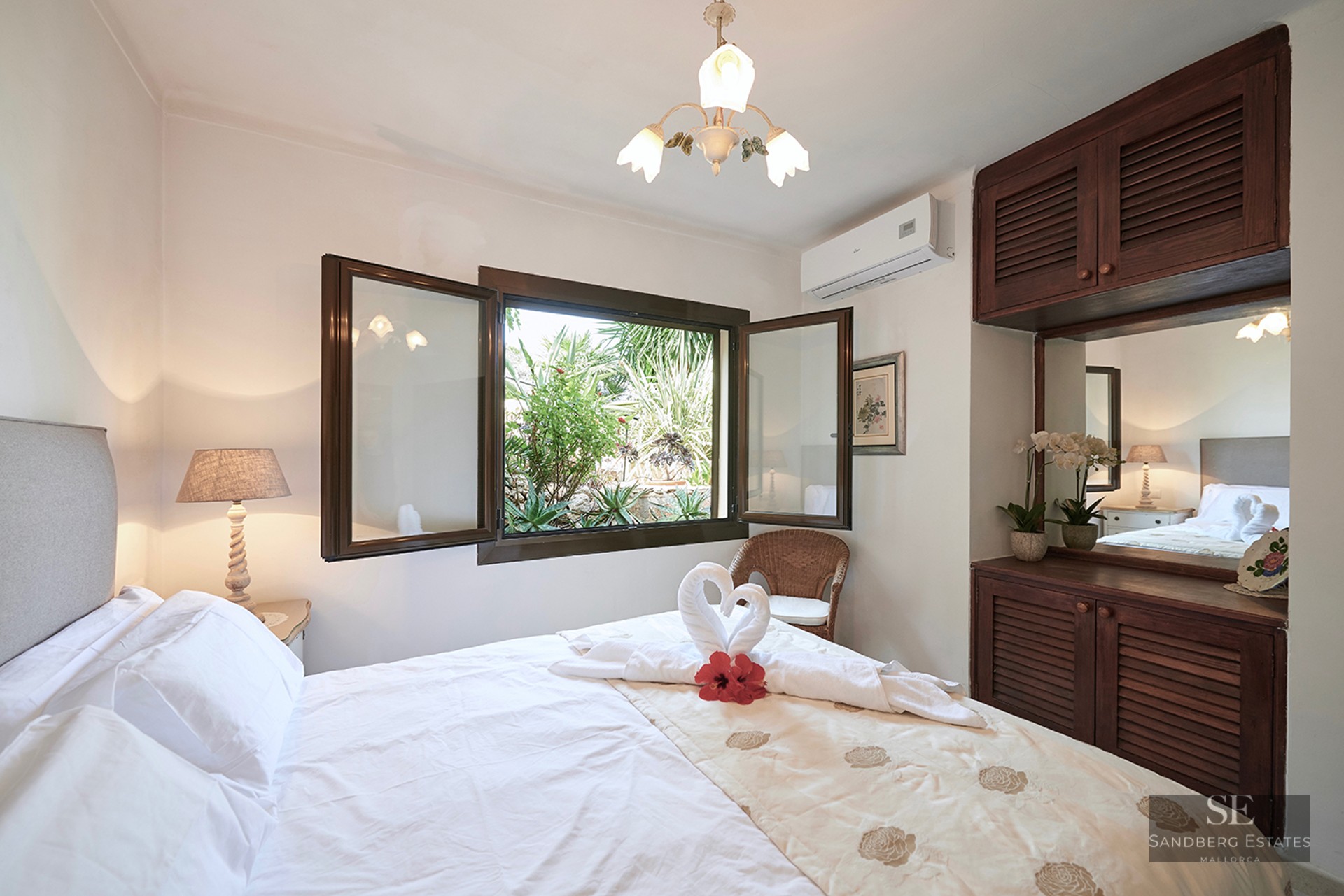 Bright bedroom with white linens, dark wood closets, and an open window looking onto a lush green garden.