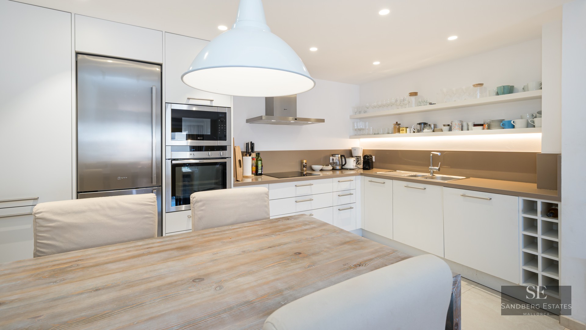 Modern white kitchen with stainless steel appliances, a wooden table, and beige upholstered chairs under a pendant light.