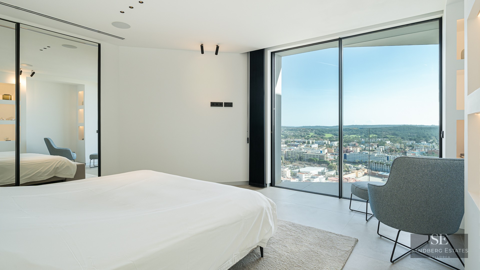 Minimalist bedroom featuring a white bed, grey armchair, and floor-to-ceiling windows overlooking a city cityscape.
