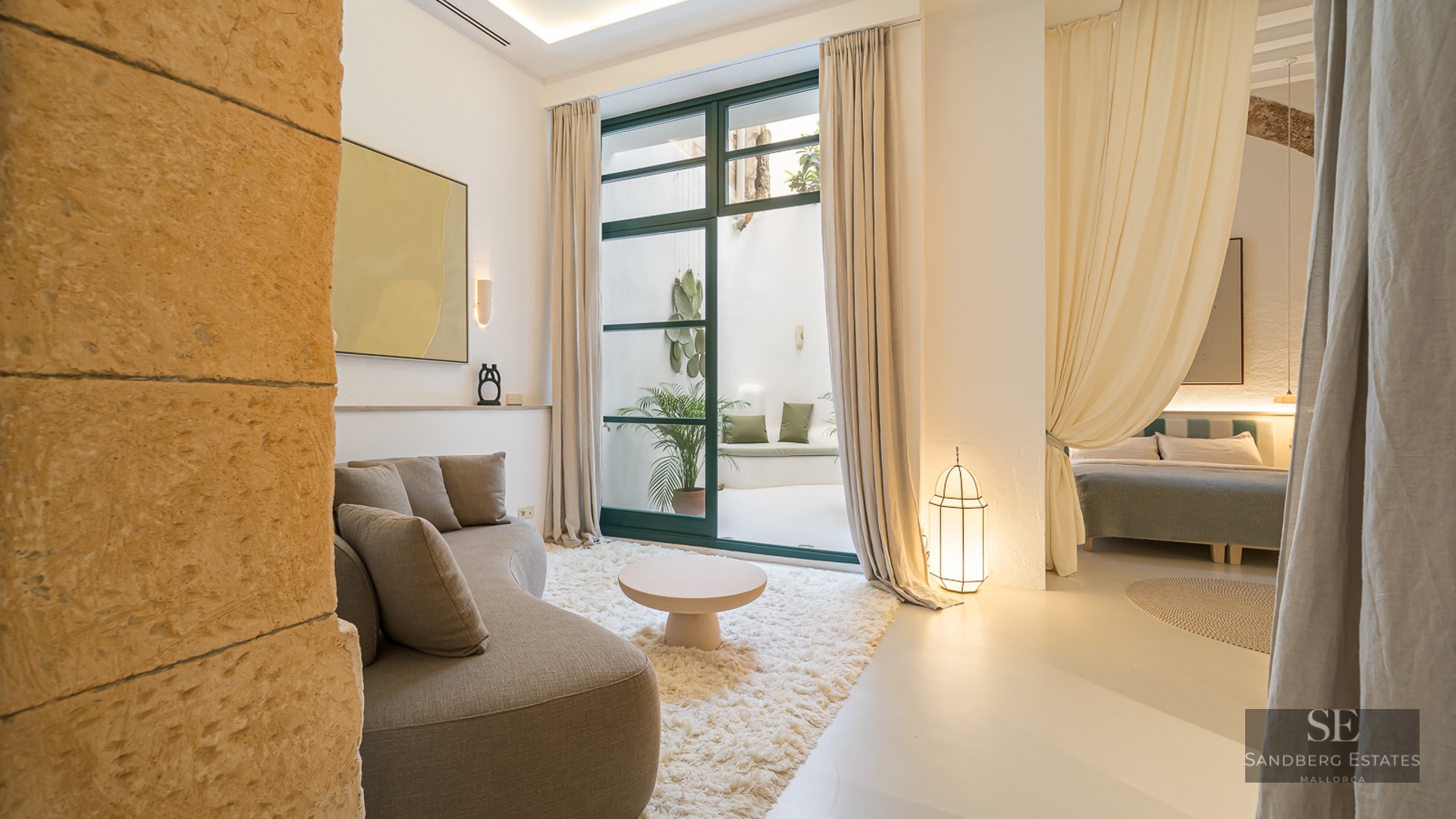 Sun-drenched living room with a curved sofa, white shag rug, stone wall, and glass doors opening to a lush patio.