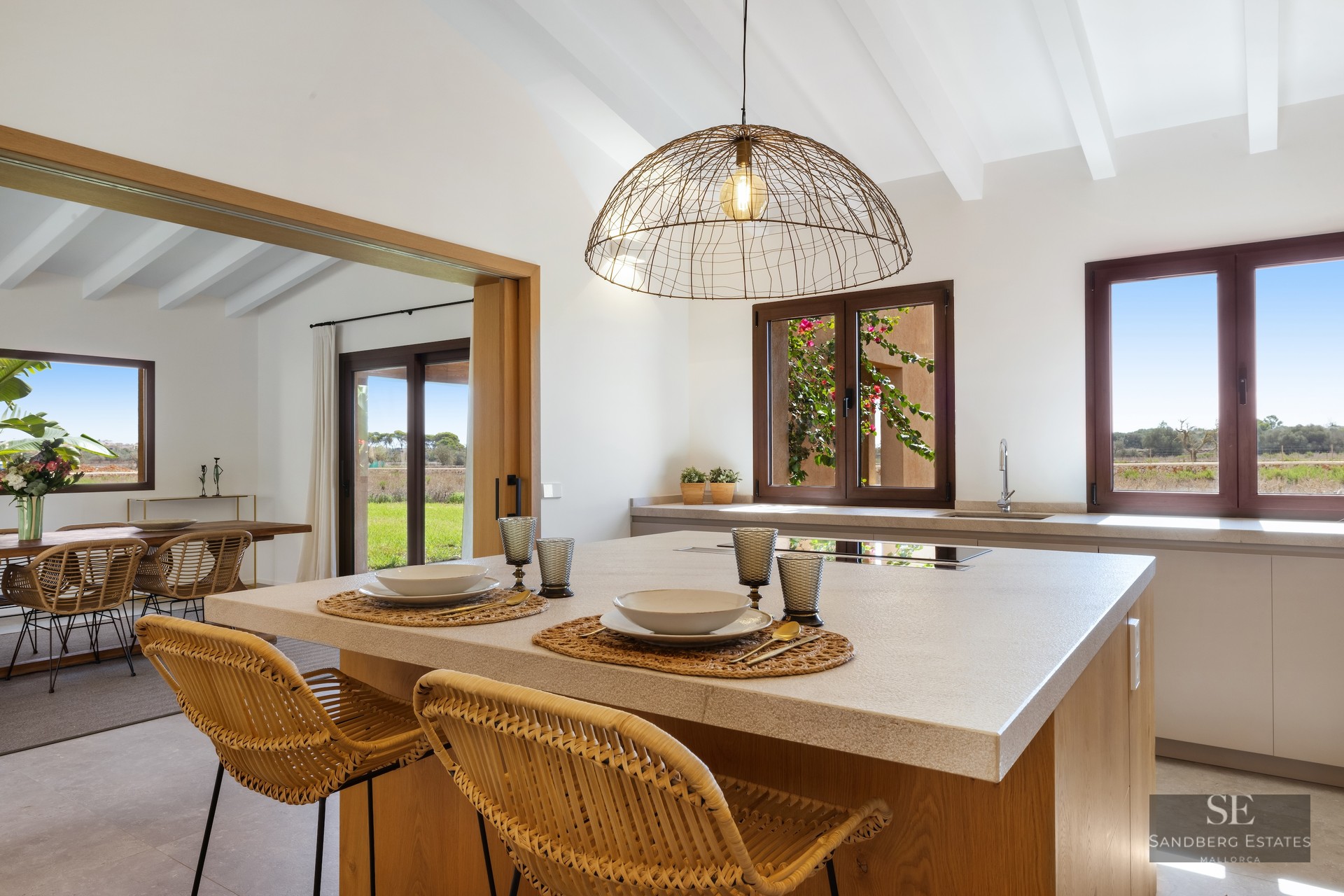 Modern kitchen island with wicker stools and white beamed ceiling overlooking rural landscape.