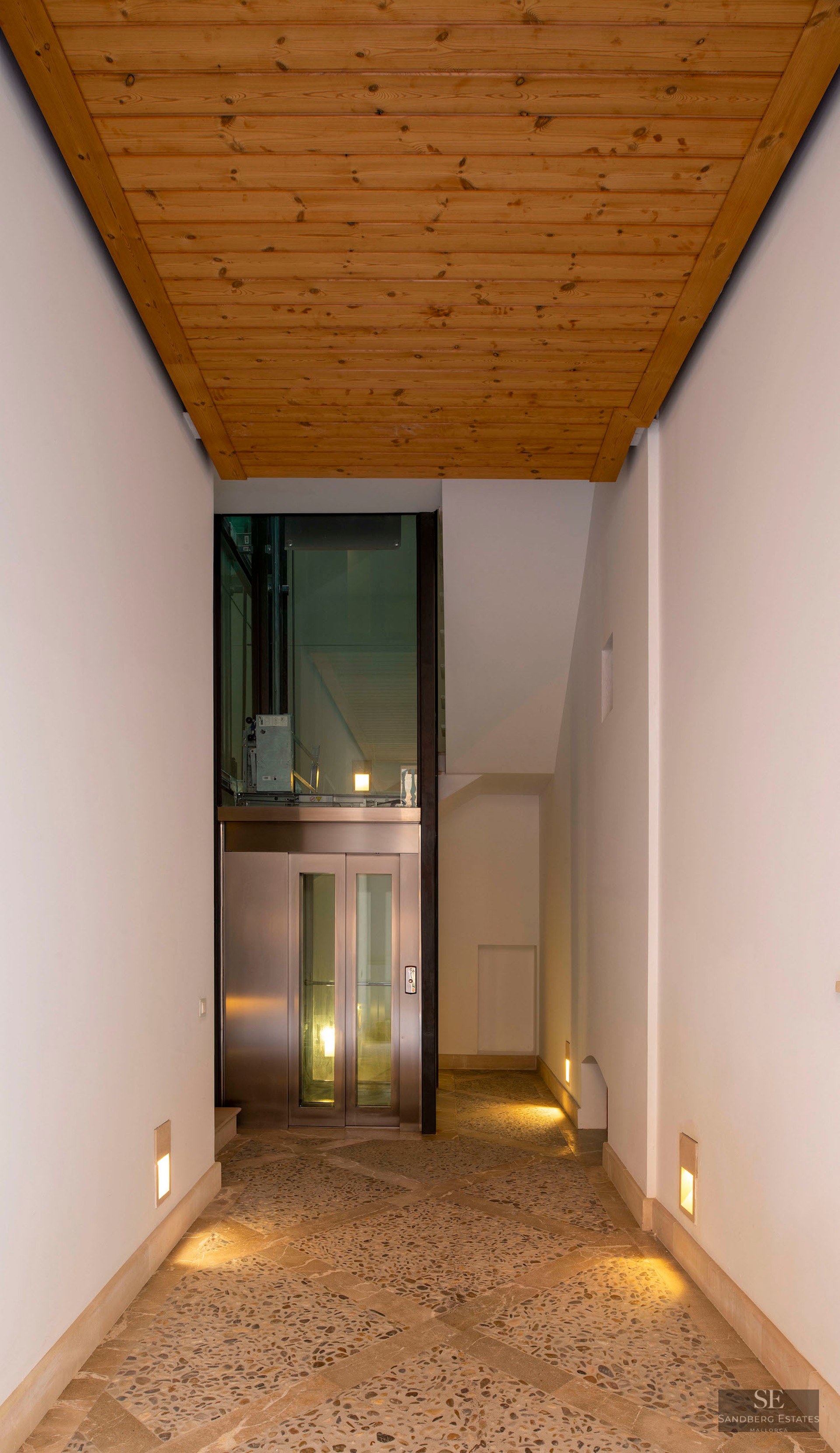 Modern hallway with stone mosaic flooring, wood ceiling, and a glass elevator at the end.