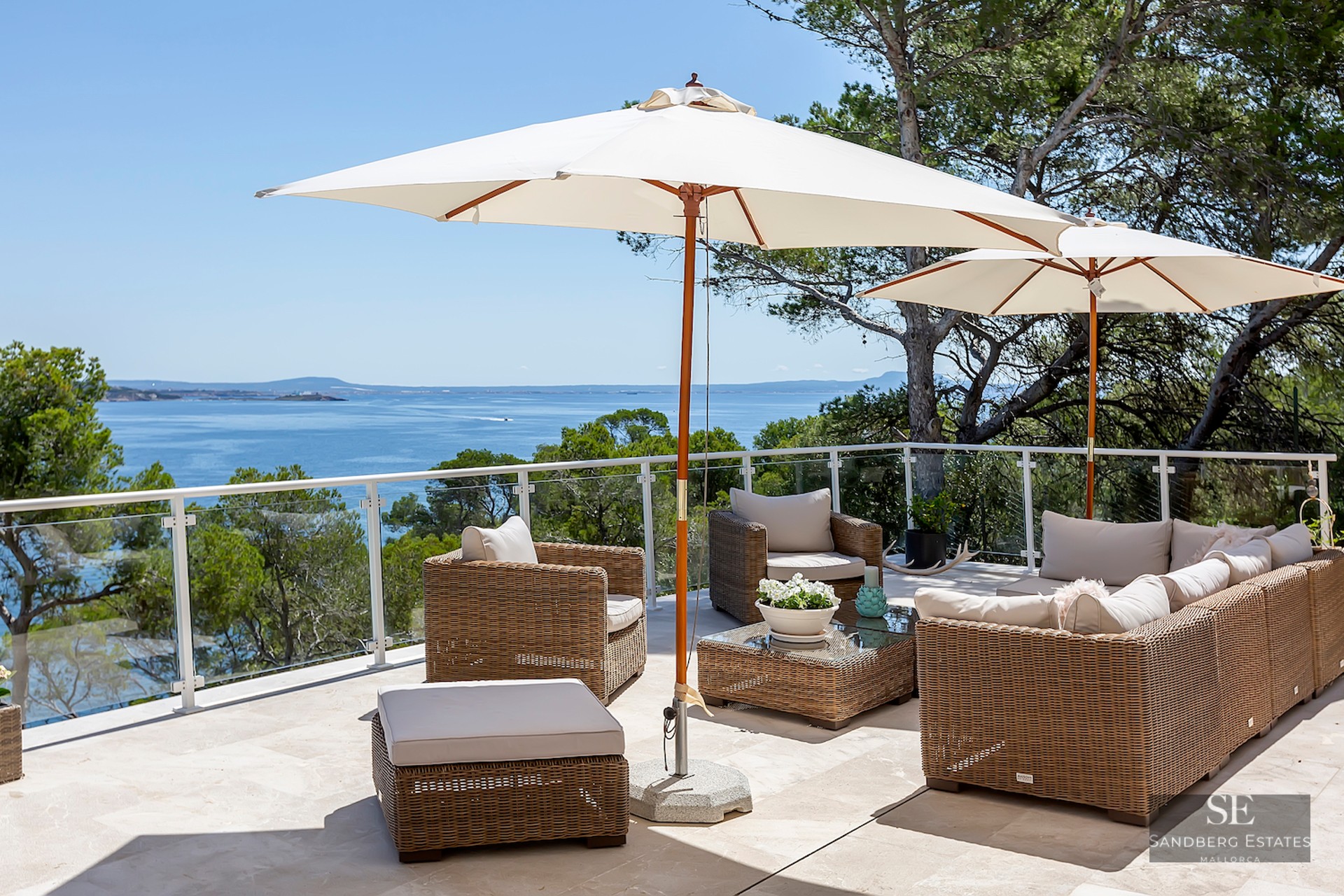 Sun-drenched terrace with wicker lounge furniture, white umbrellas, and glass railings overlooking the blue sea.
