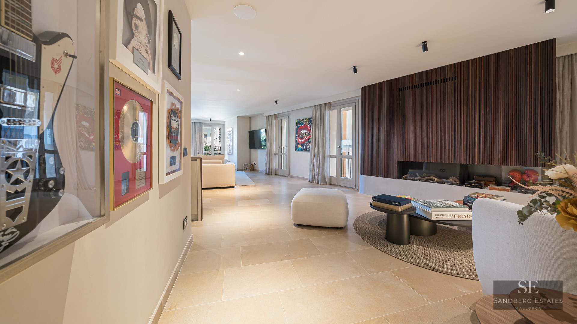 Modern living room featuring a wood-slat fireplace wall, stone floors, and framed music memorabilia in a hallway.