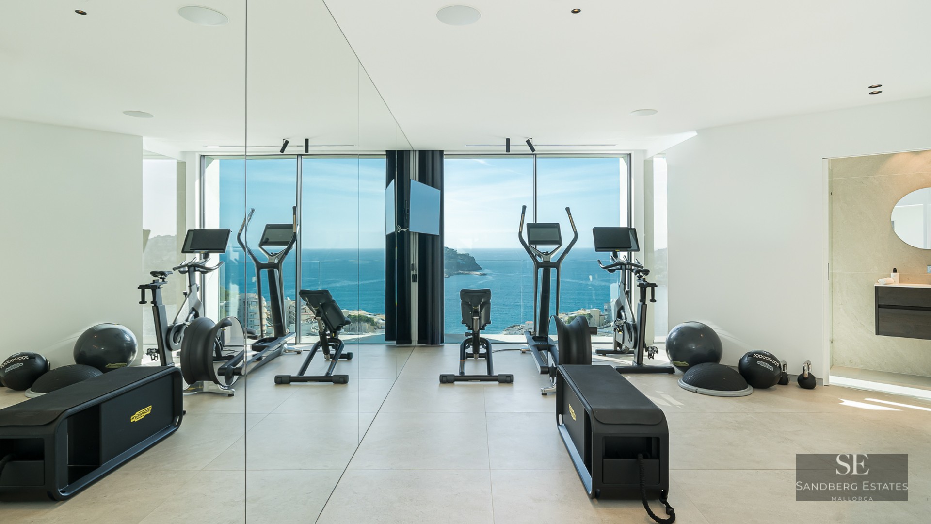 Modern home gym with professional equipment and floor-to-ceiling windows overlooking the blue Mediterranean sea.