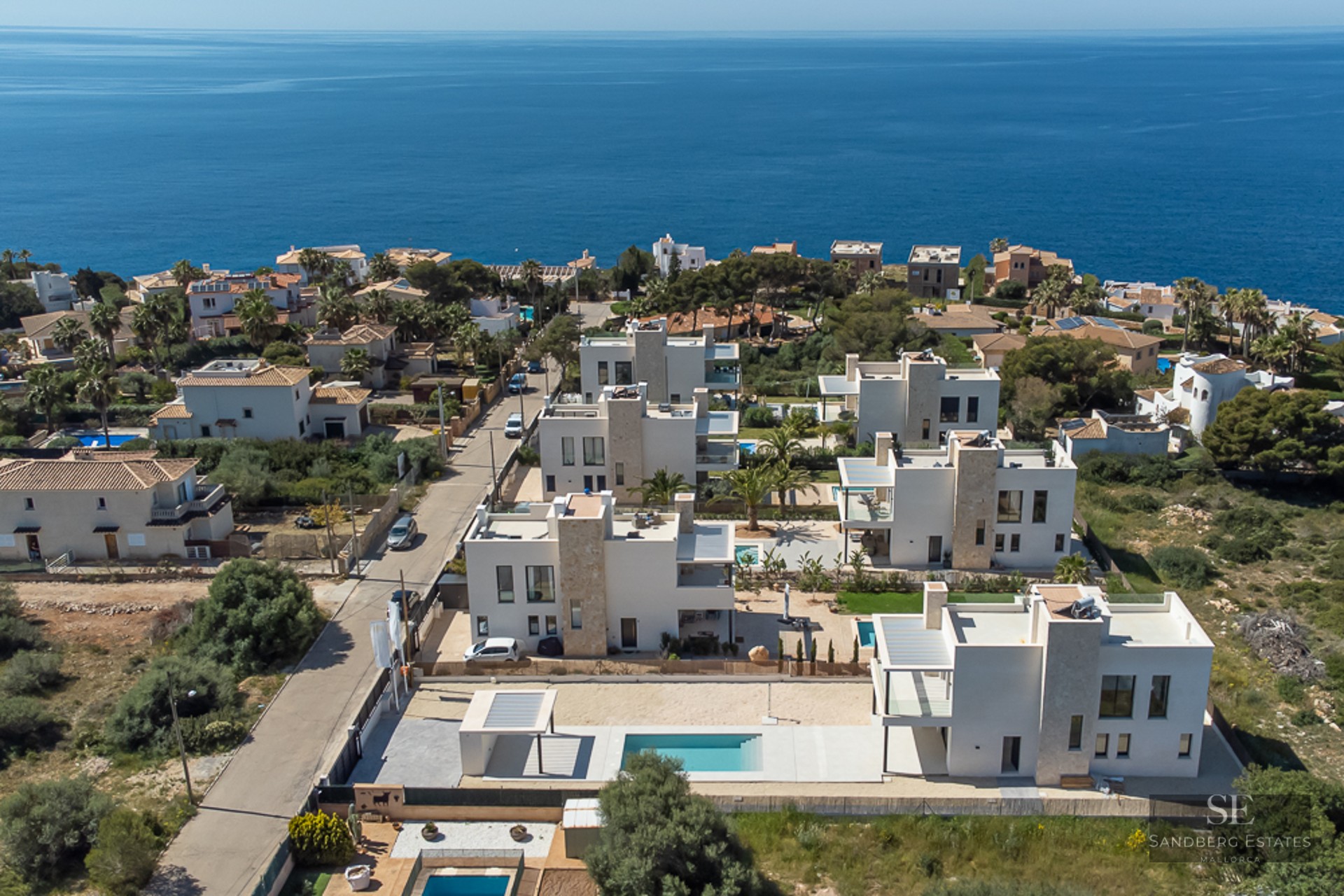 Aerial view of a cluster of modern white villas with private pools overlooking the blue Mediterranean Sea.