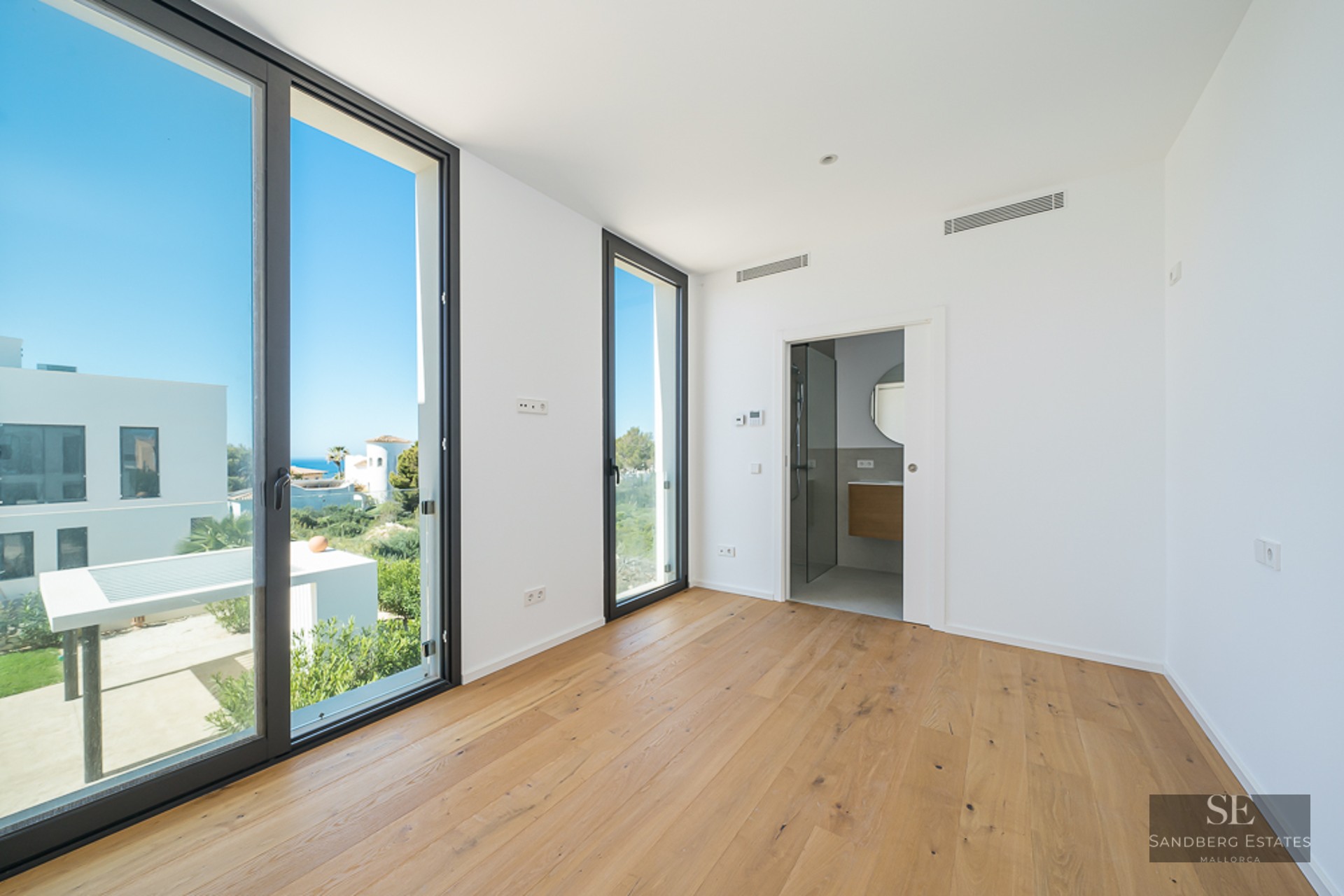 Bright empty bedroom with light wood floors, large floor-to-ceiling windows, and access to an ensuite bathroom.