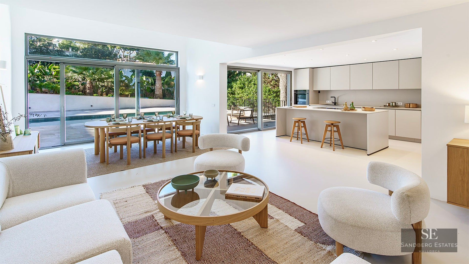 Bright open-plan room with modern white sofas, wood dining set, and minimalist kitchen overlooking a pool and garden.