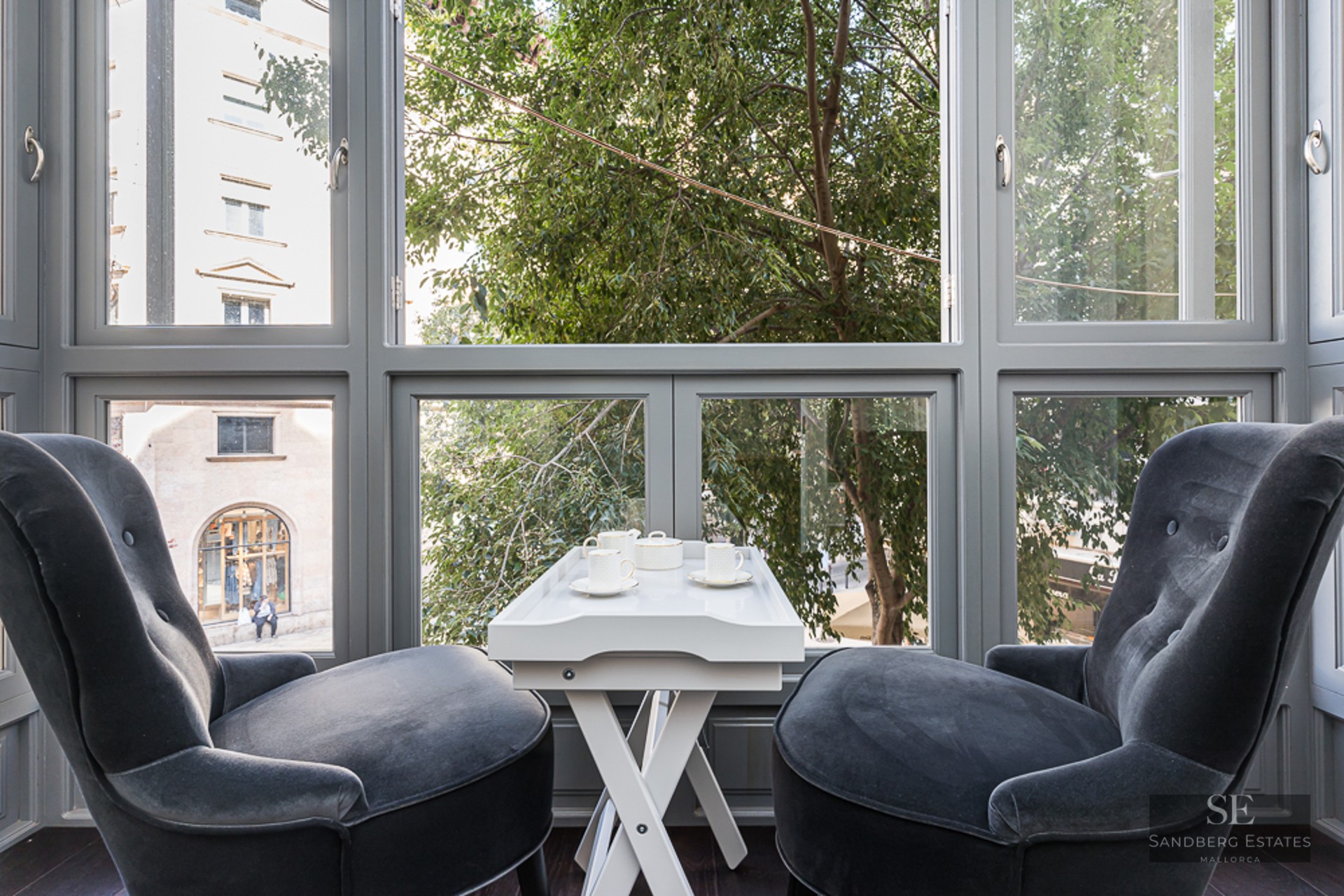 Two charcoal velvet armchairs facing a large window with a white tea table in between, overlooking city trees.