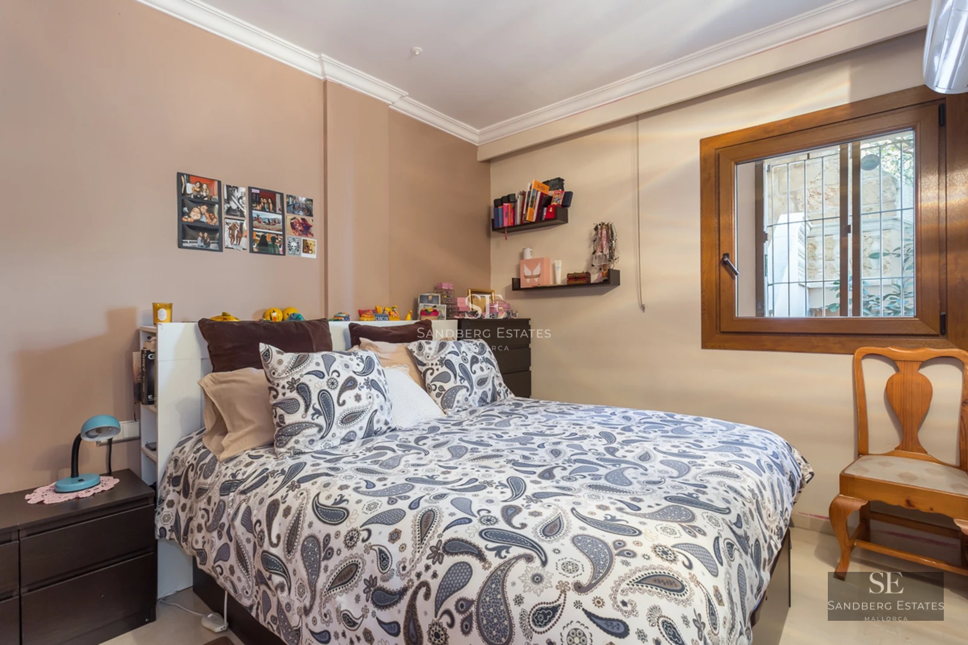 A bedroom featuring a paisley-patterned bed, taupe walls, a wooden window frame, and a vintage-style wooden chair.
