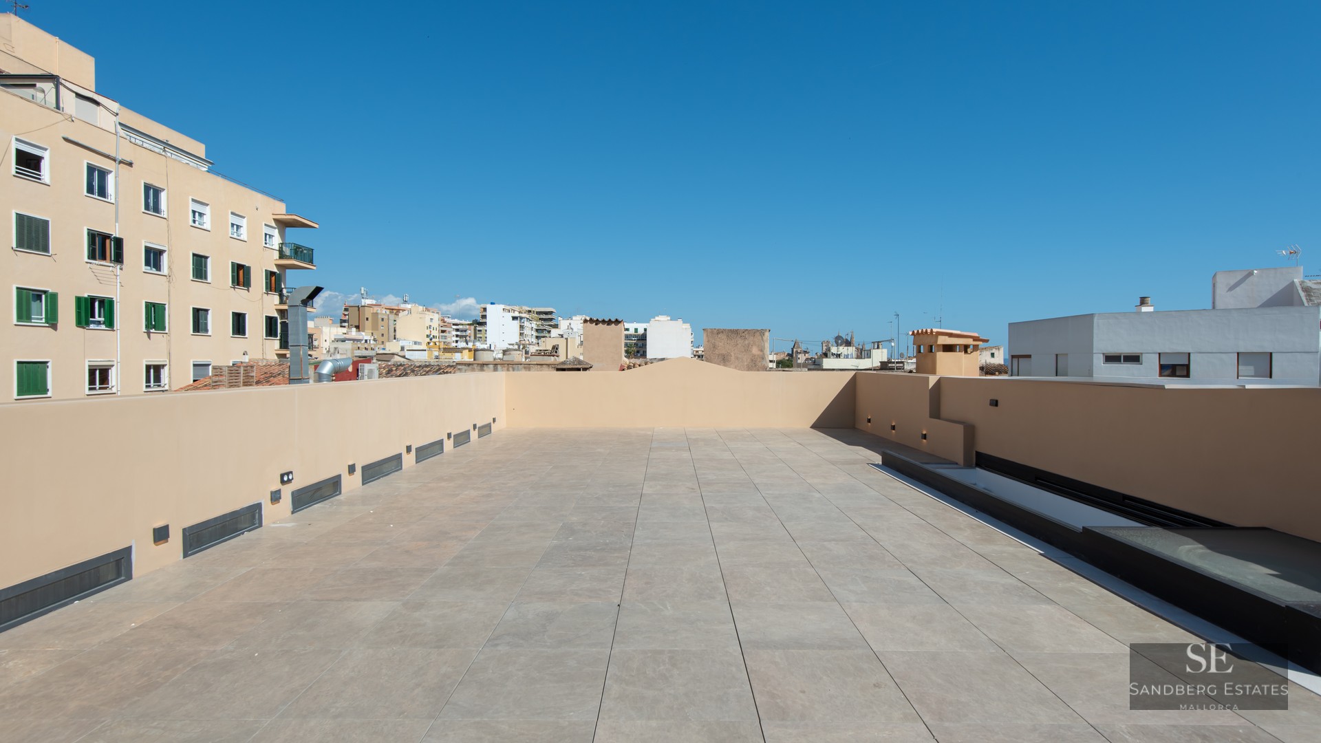 Spacious rooftop terrace with beige tiles and modern wall lighting under a clear blue sky.