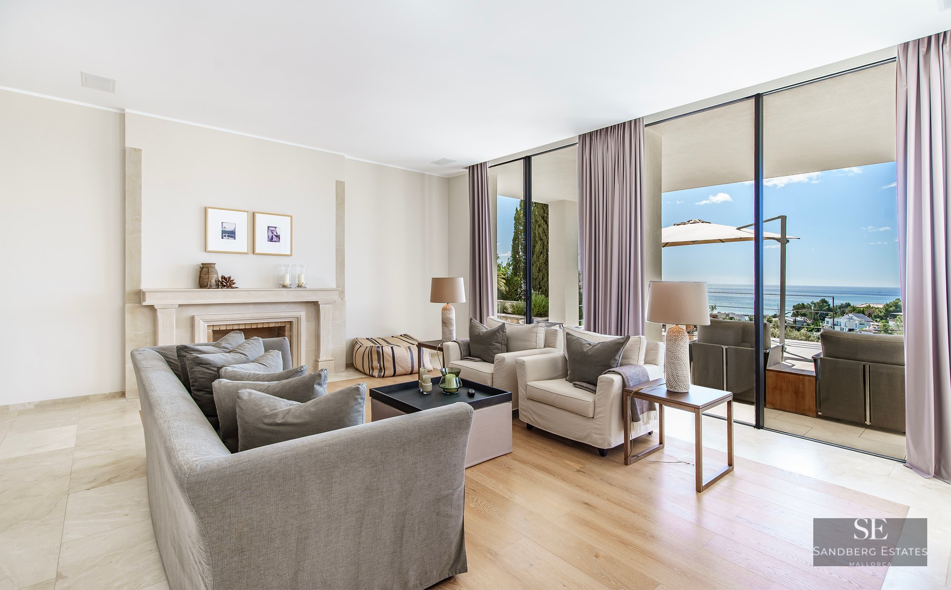 Modern living room with grey sofas, a stone fireplace, and floor-to-ceiling windows opening to a sea-view terrace.