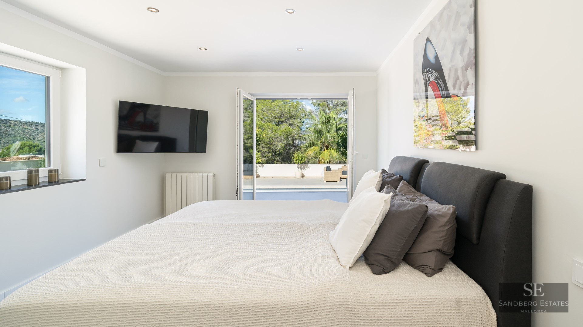 Chambre moderne et lumineuse avec un grand lit, TV murale et portes-fenêtres ouvrant sur une terrasse ensoleillée.