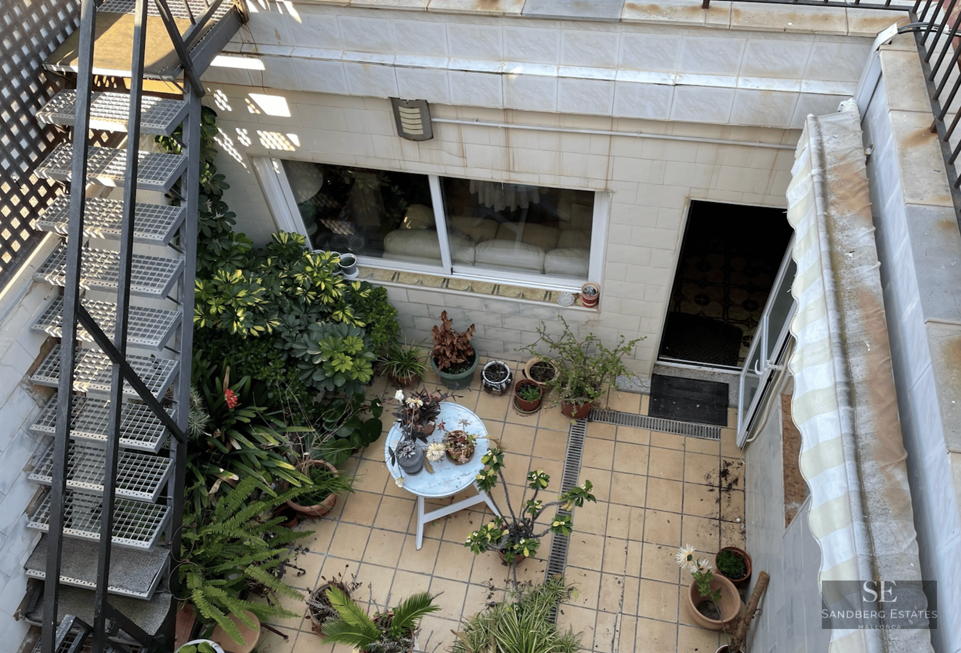 Overhead view of a tiled patio with a metal staircase, numerous potted plants, and a small white round table.