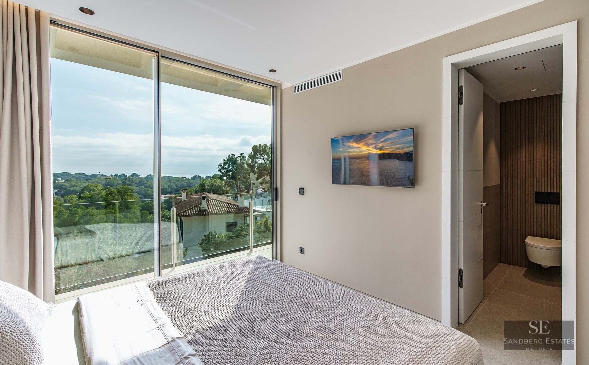 A modern bedroom with a large floor-to-ceiling window overlooking a forest and sea, with an ensuite bathroom visible.