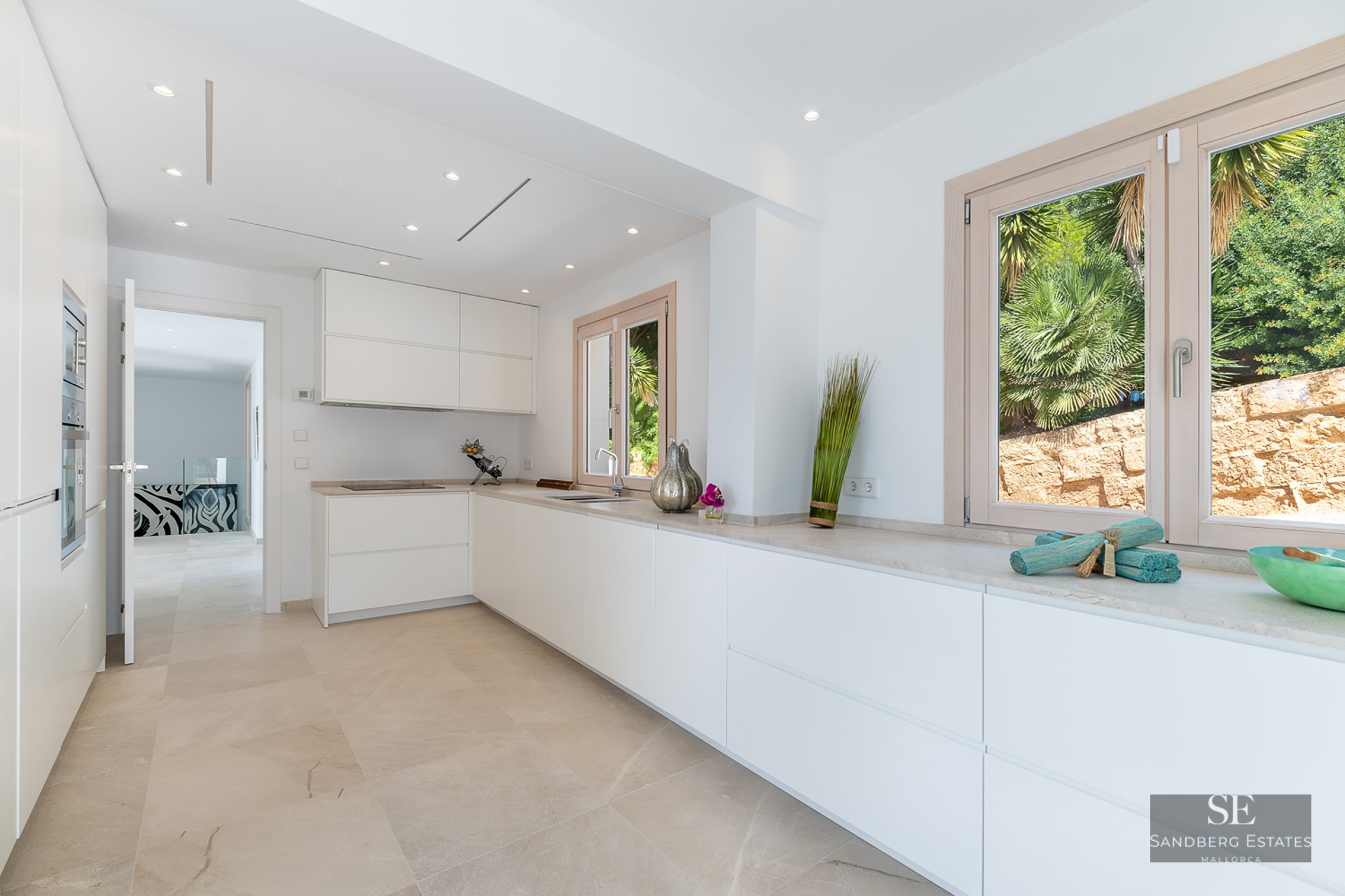 Bright, modern kitchen featuring white handleless cabinets, stone countertops, and windows overlooking greenery.