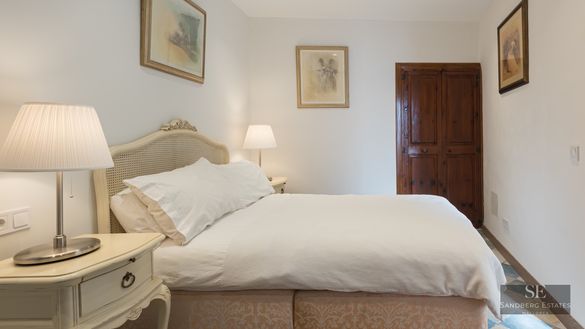 A cozy bedroom featuring a white bed with a cane headboard, twin bedside lamps, and a dark vintage wooden door.