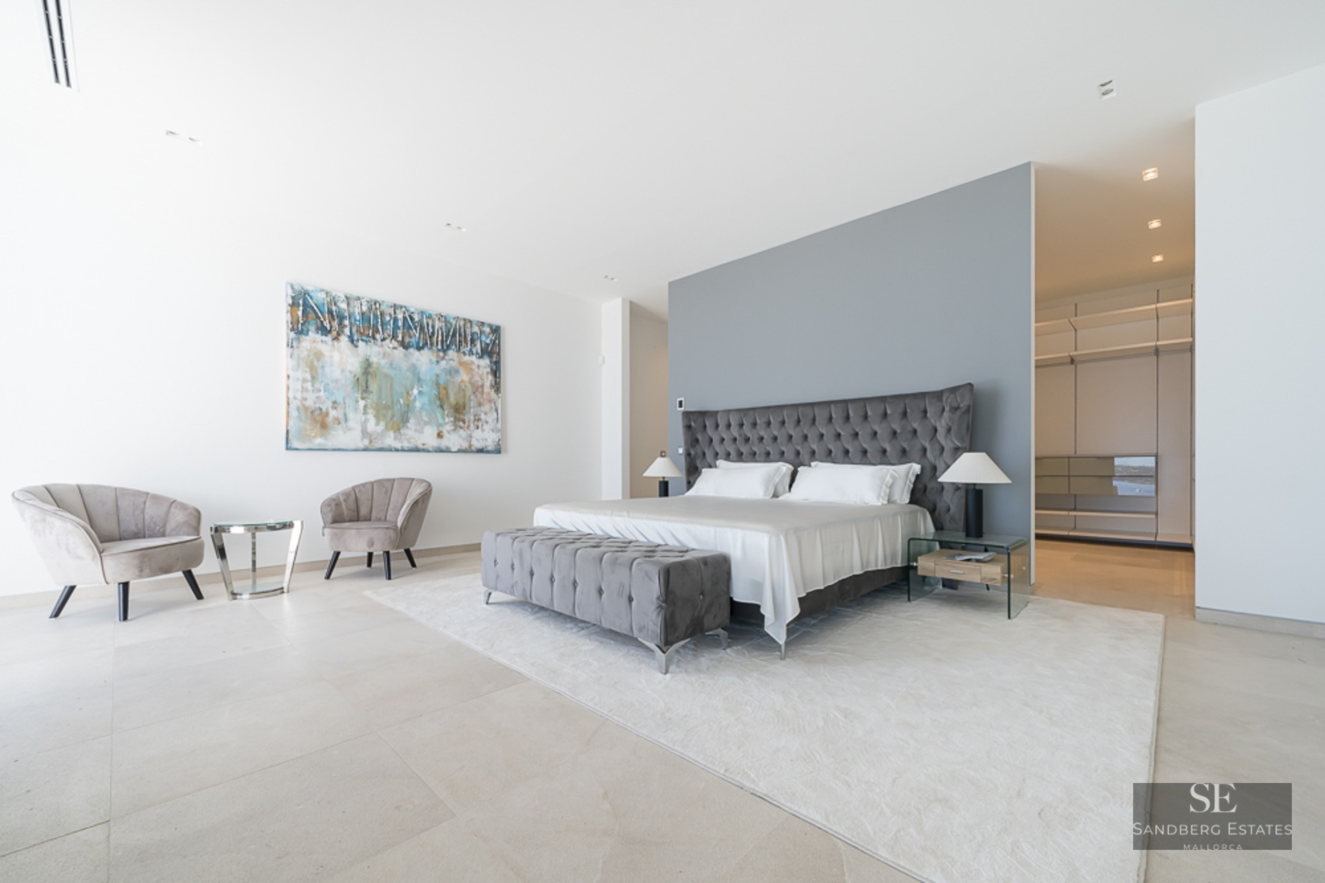 Master bedroom featuring a grey tufted bed, velvet seating area, and a walk-in wardrobe in the background.