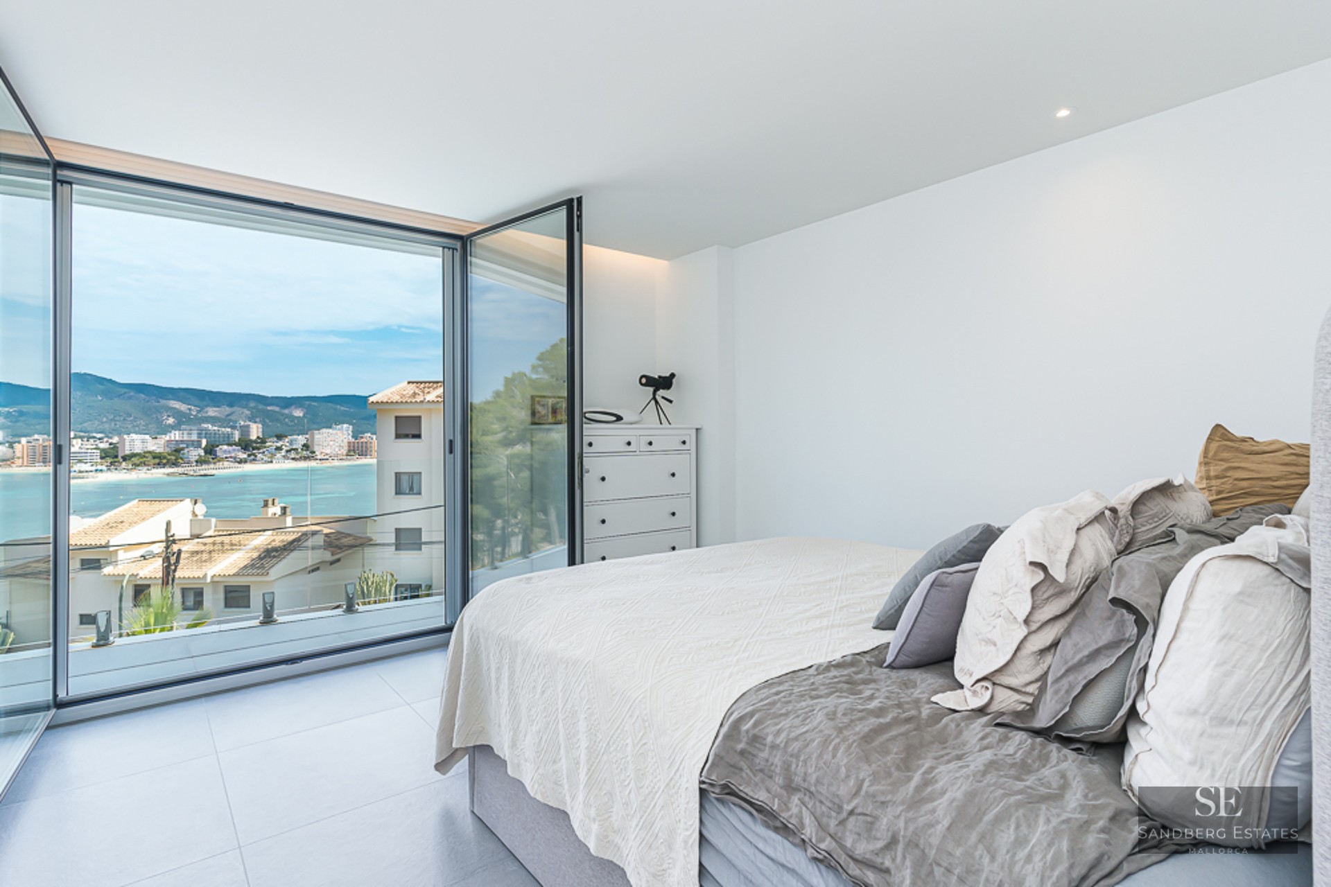 Bright bedroom with a bed and large glass doors opening to a coastal bay view and mountains.