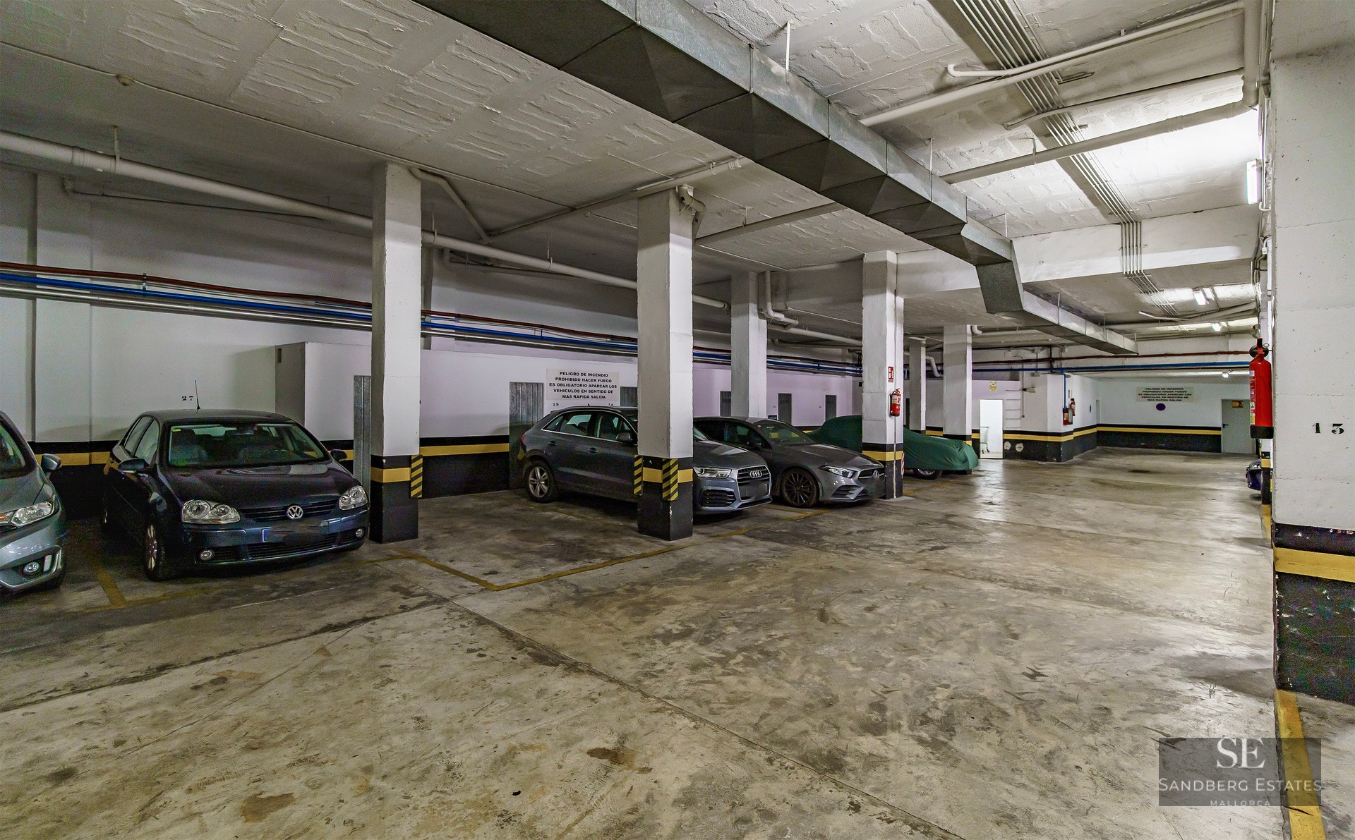 Grand garage souterrain avec piliers en béton, marquages de sécurité jaunes et voitures garées.