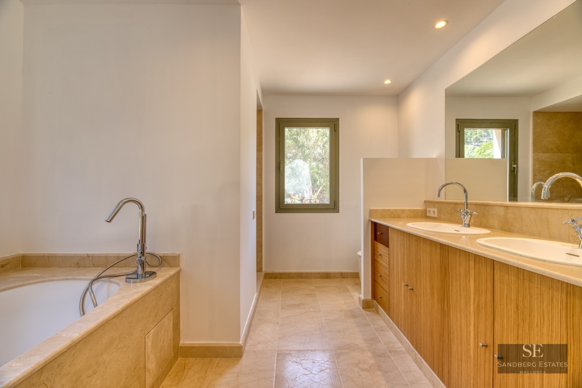 Luxury bathroom featuring wooden double vanity, beige marble countertops, bathtub, and natural light.