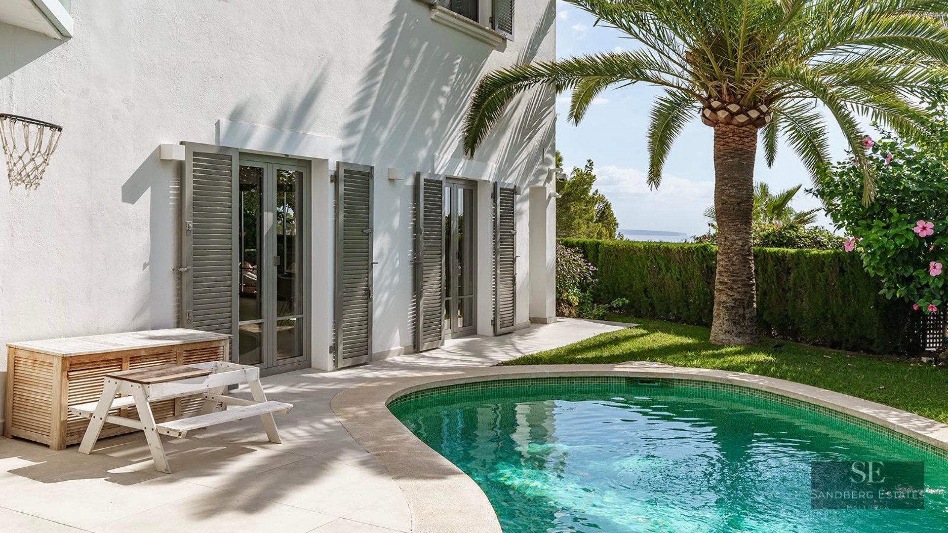A turquoise swimming pool beside a white villa with a palm tree, grey shutters, and a distant ocean view.