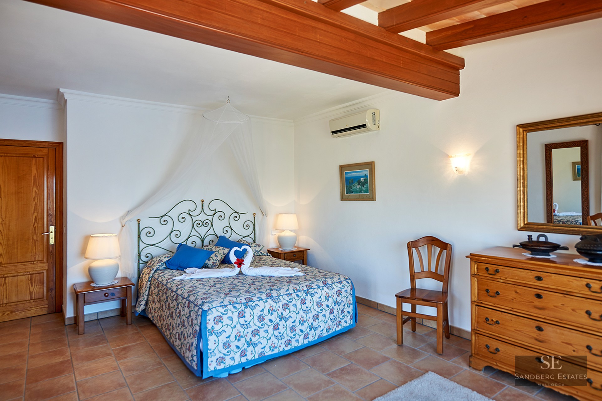Bedroom with terracotta tile floor, exposed wooden ceiling beams, wrought iron bed, and classic wooden furniture.