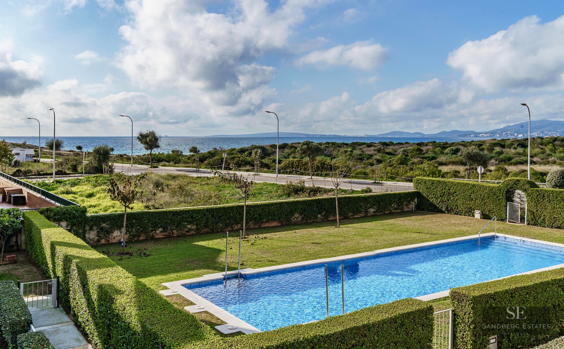 Rectangular blue pool surrounded by green lawn and hedges with the Mediterranean sea in the background under cloudy sky.